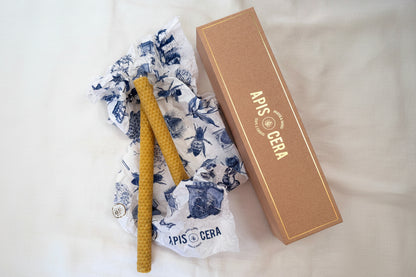 Deux bougies Lucienne en cire d'abeille gaufrée avec leur boîte et papier d'emballage illustré.